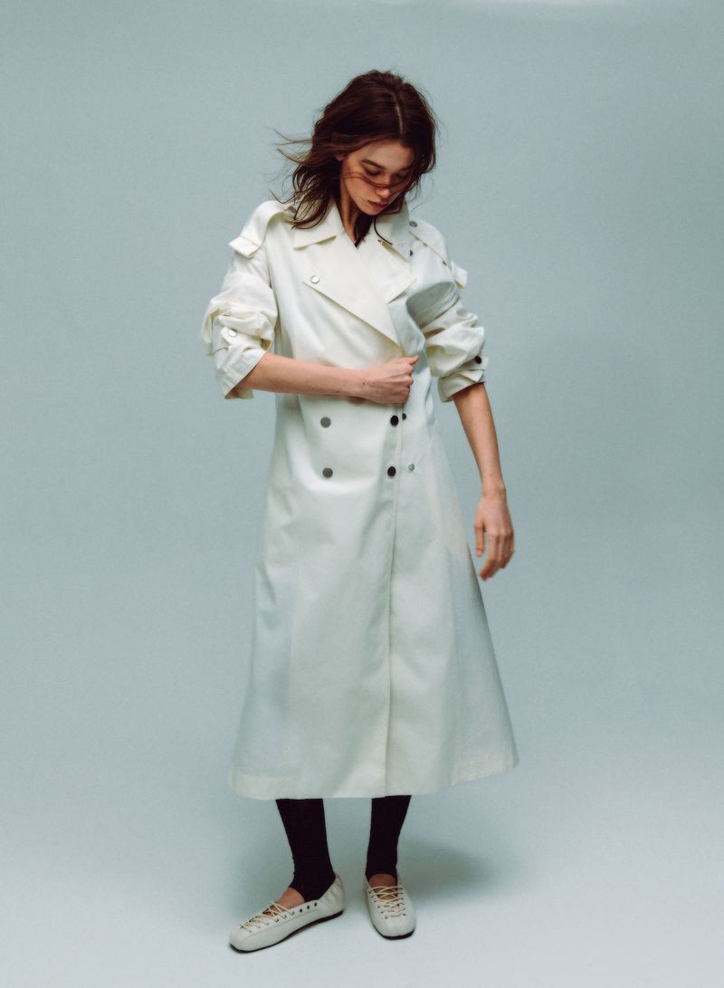 Model wearing a long white trench coat and light shoes against a plain grey studio background.
