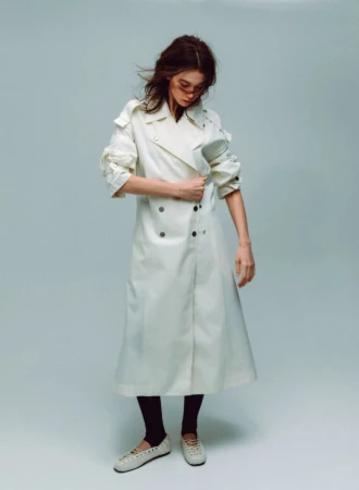 Model wearing a long white trench coat and light shoes against a plain grey studio background.