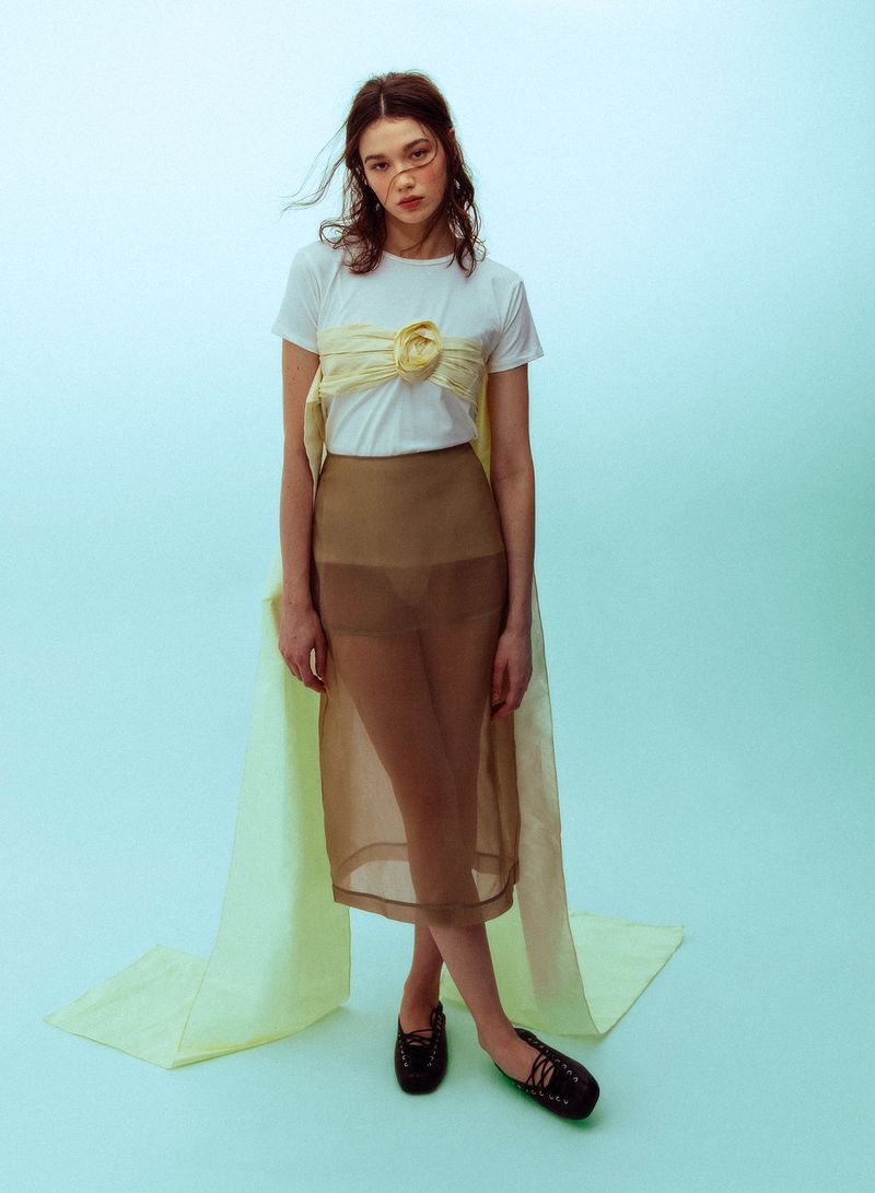 Model wearing a white t-shirt, sheer brown skirt, and pale yellow sash against a light blue seamless backdrop