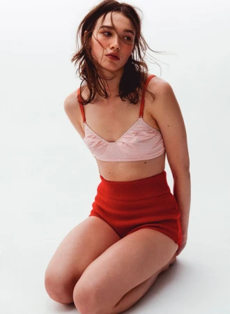 Model wearing a pink bralette and red high-waisted shorts kneeling against a white studio background.