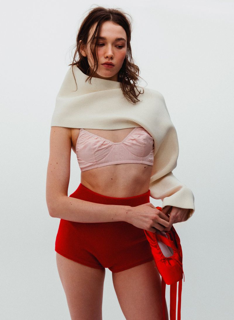 Model in pink bralette, red shorts and cream wrap scarf holding red pointe shoes against a white background