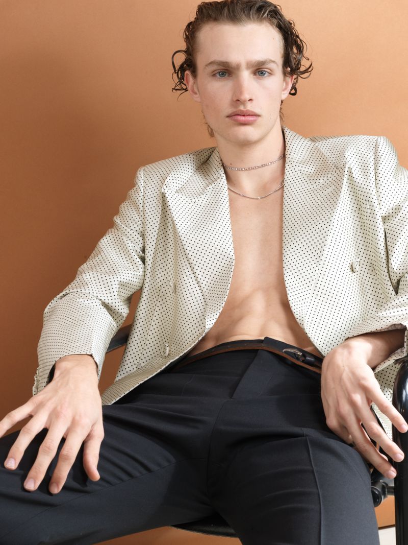 A male model seated wearing an off-white patterned blazer and dark trousers against a warm studio backdrop.