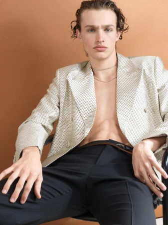 A male model seated wearing an off-white patterned blazer and dark trousers against a warm studio backdrop.