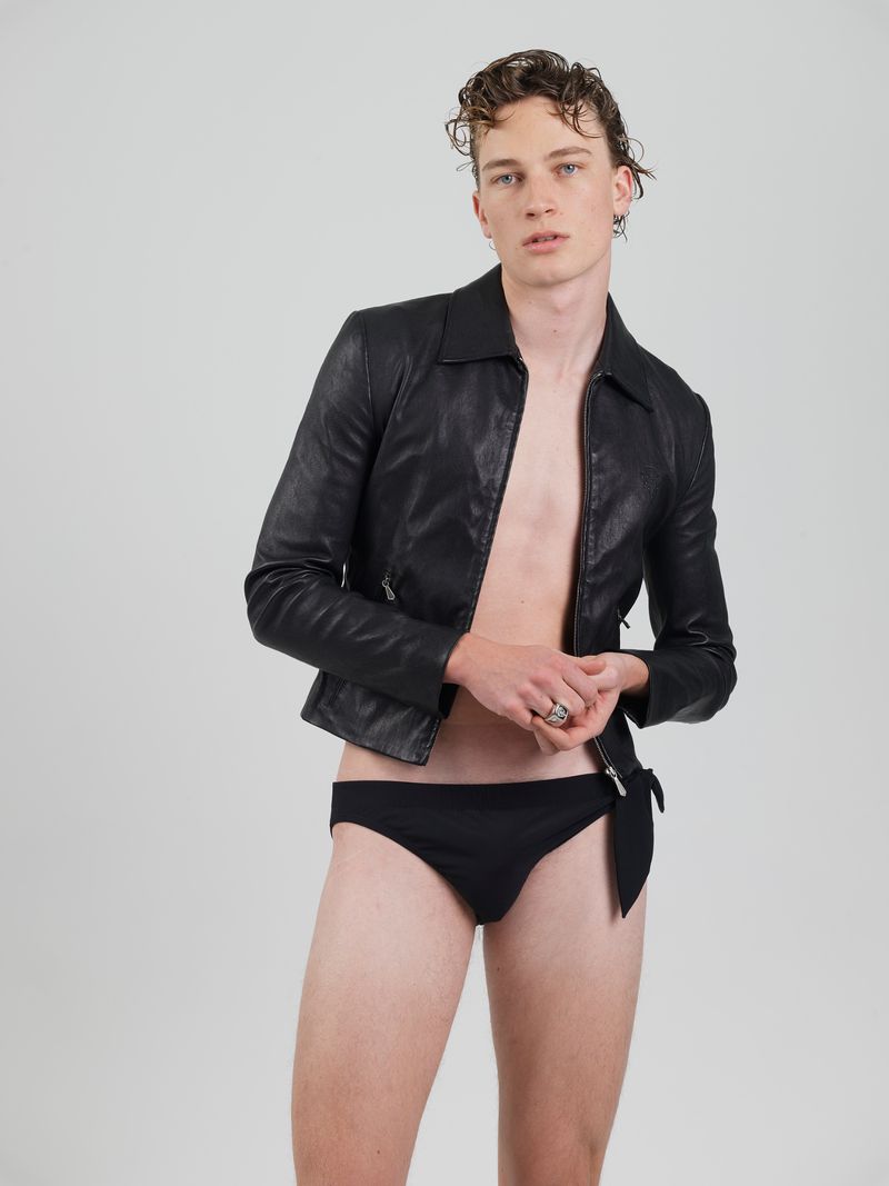 Male model wearing an unzipped black leather jacket and black briefs against a neutral light grey background.