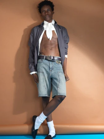Young Black male model wearing denim shorts and white socks against a warm brown backdrop