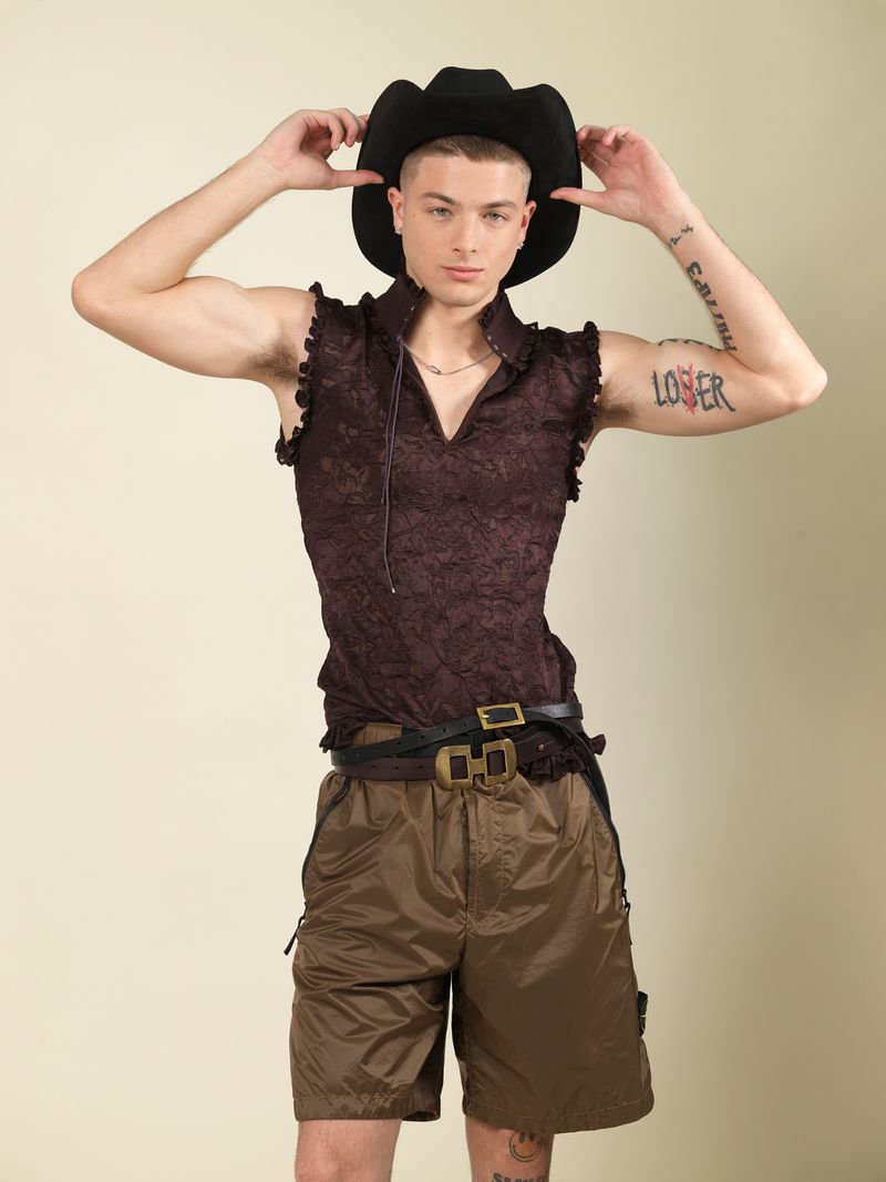 A man wearing a black cowboy hat, dark sleeveless patterned shirt, and brown shorts standing against a light background