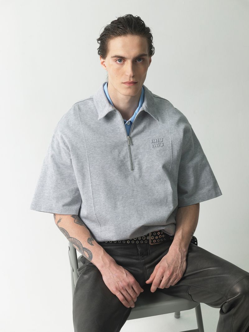 Adult male with tattoo on arm poses in gray, half-zip shirt and black pants while seated on a metal chair in a photo studio.