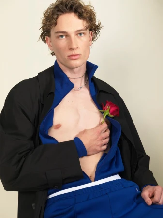 Young male model holding a red rose against his chest, wearing a black jacket and blue zip-up top.