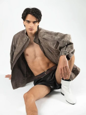 A male model seated on a white background wearing an oversized brown jacket, dark shorts, and white boots.