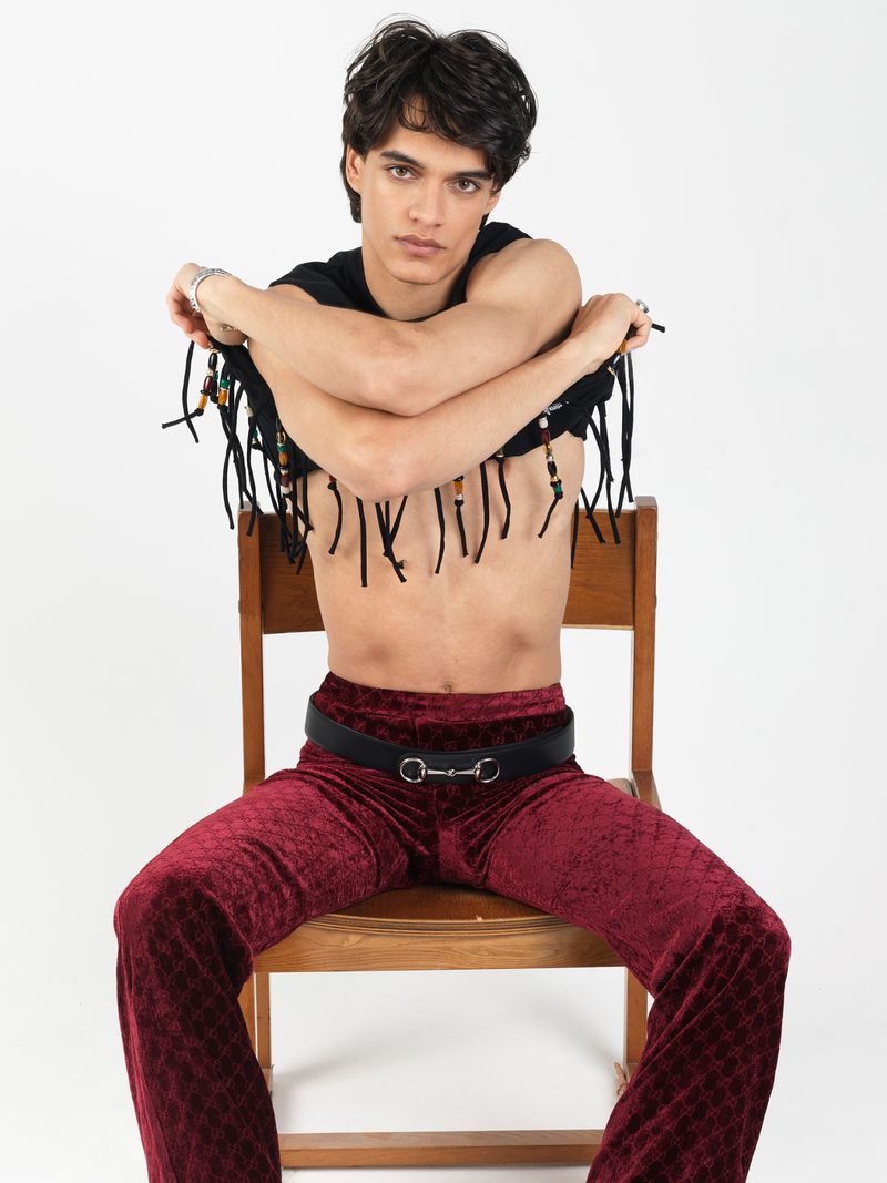 Young man with dark hair wearing fringed black top and red velvet pants seated on a wooden chair against a plain white background