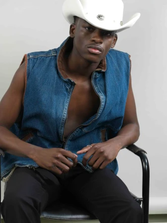 A Black man wearing a white cowboy hat and blue denim vest seated in a chair against a neutral background.