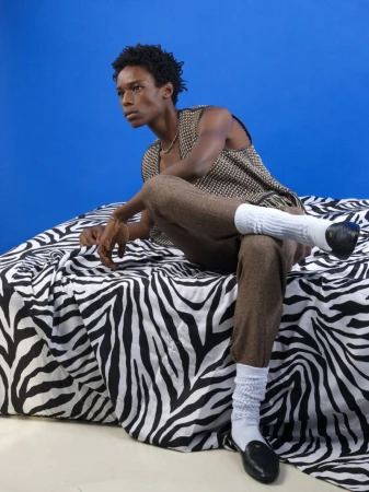 Young Black male model seated on a zebra-print surface against a bright blue backdrop wearing patterned vest and brown trousers