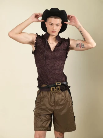 A man wearing a black cowboy hat, dark sleeveless patterned shirt, and brown shorts standing against a light background