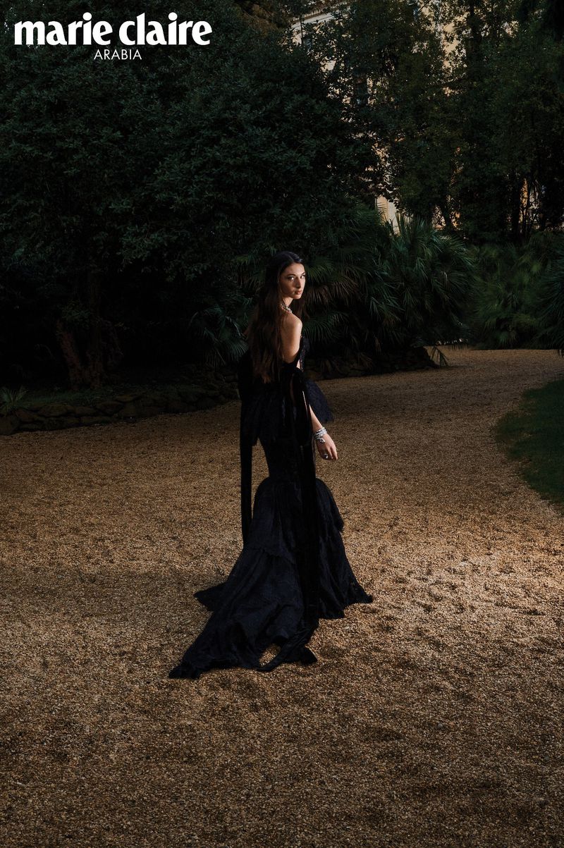 Model wearing a black evening gown walking along a gravel park path surrounded by dark dense foliage