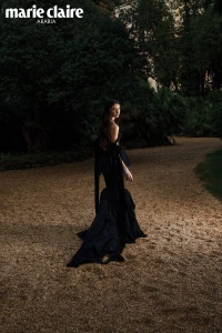 Model wearing a black evening gown walking along a gravel park path surrounded by dark dense foliage