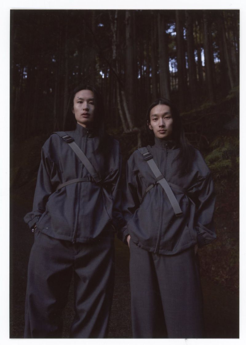 Two slender Asian women in matching athleisure wear pose in a dimly lit forest, wearing crossbody bags and long dark hair.