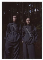 Two models wearing dark technical jackets with diagonal straps standing outdoors in a dense forest setting.