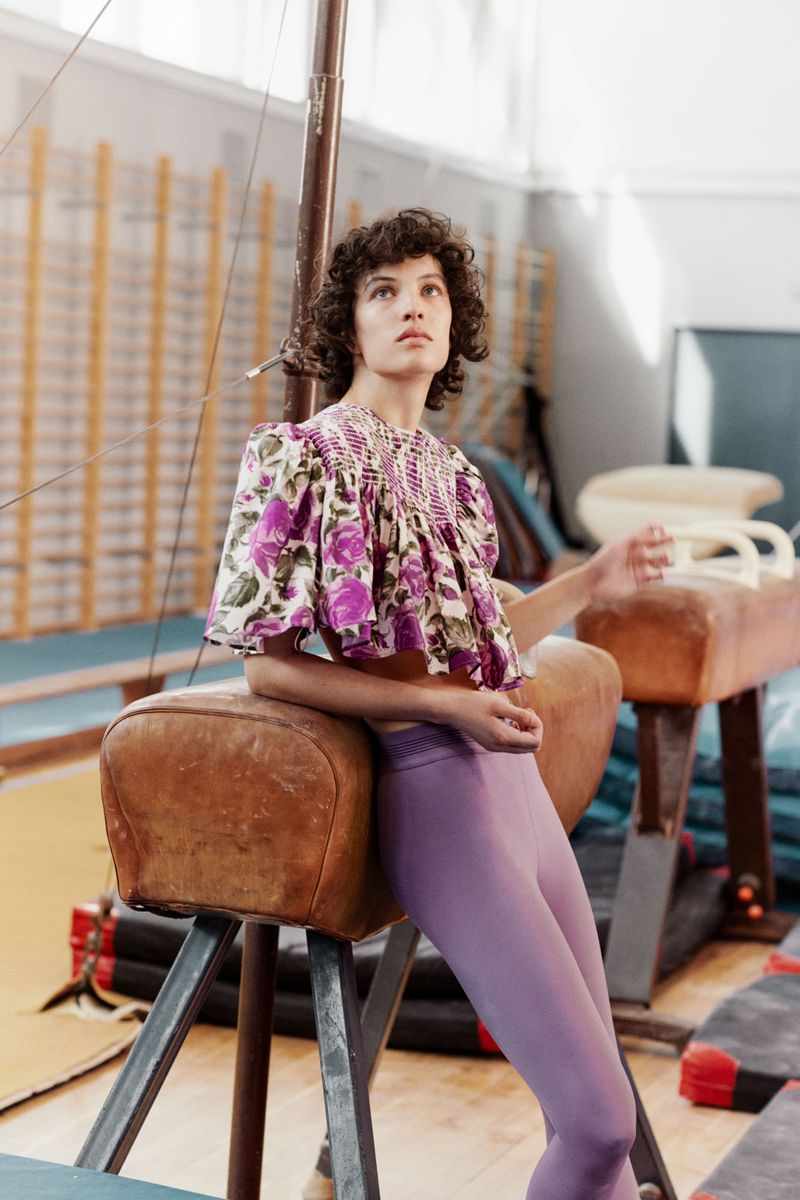A woman in a floral top and lavender leggings rests on a vintage gymnastic vault in a gymnasium with natural light and a contemplative mood.