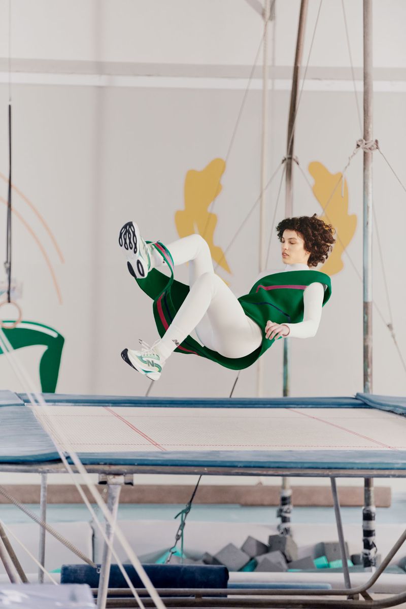 Model with curly hair in a green vest and white leggings mid-air above a trampoline in a gym