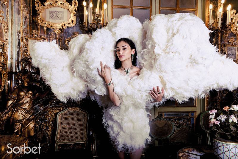 Woman wearing an extravagant white feathered haute couture dress framed by an ornate historical interior with gilded furnishings.