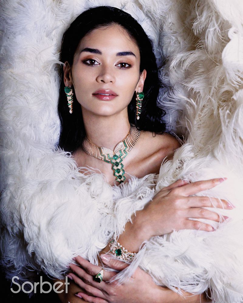 Close-up portrait of a woman surrounded by white feathers wearing emerald and diamond statement jewellery for Sorbet Magazine.
