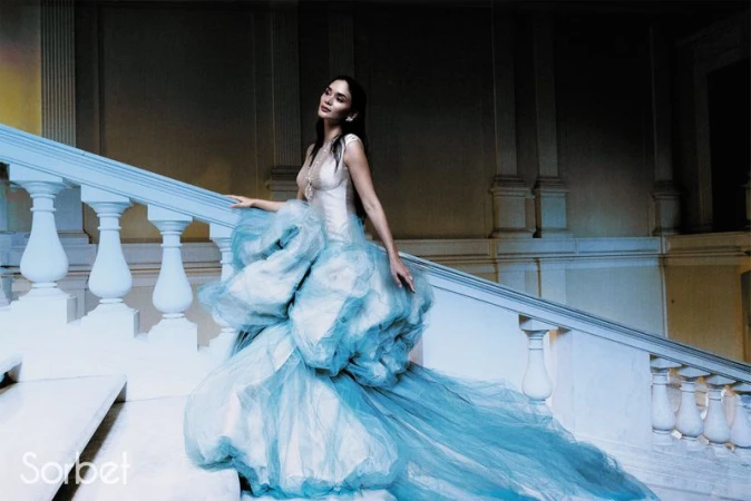 Woman in an elaborate light blue gown standing on a grand staircase inside a classical building interior