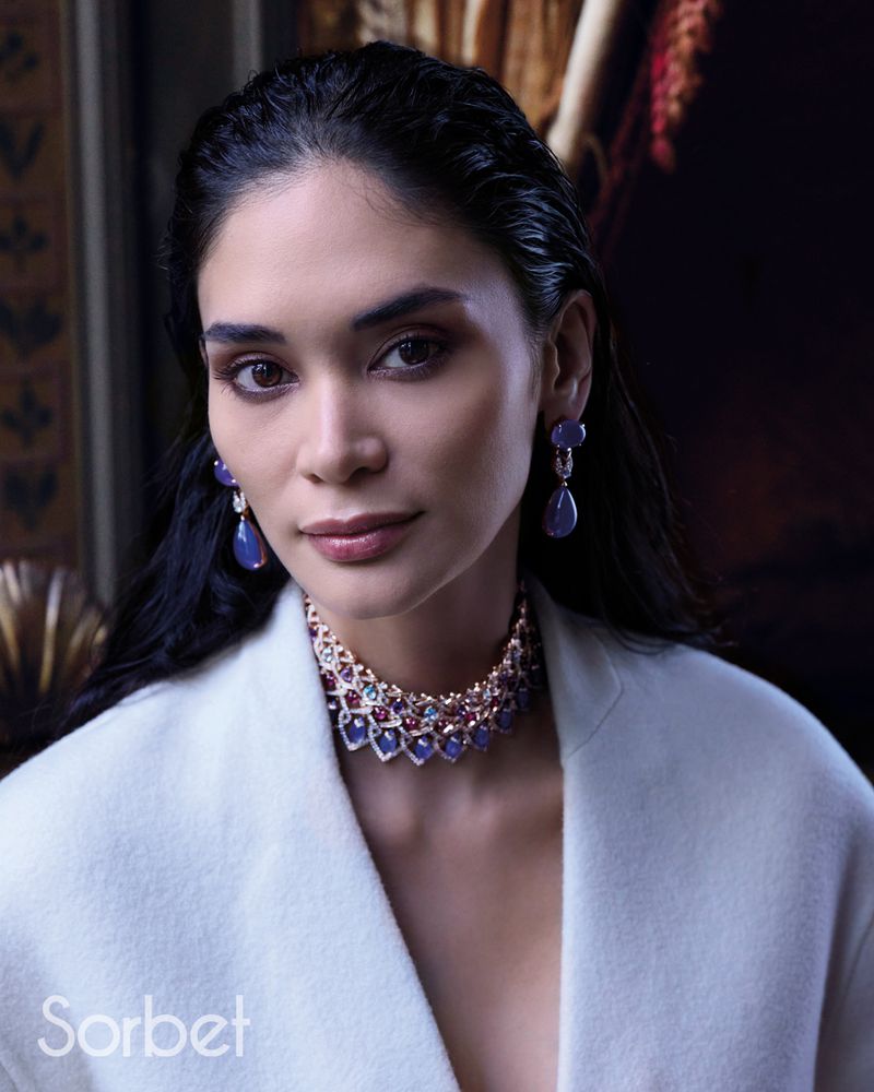 Portrait of a woman wearing a white blazer, jewel-encrusted choker necklace, and matching drop earrings against a dark background.
