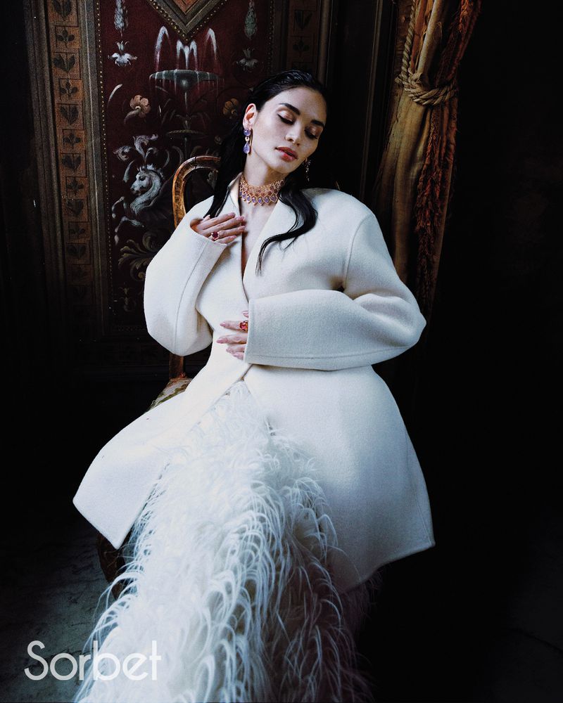 Woman wearing white structured jacket and feather skirt sitting against ornate paneling