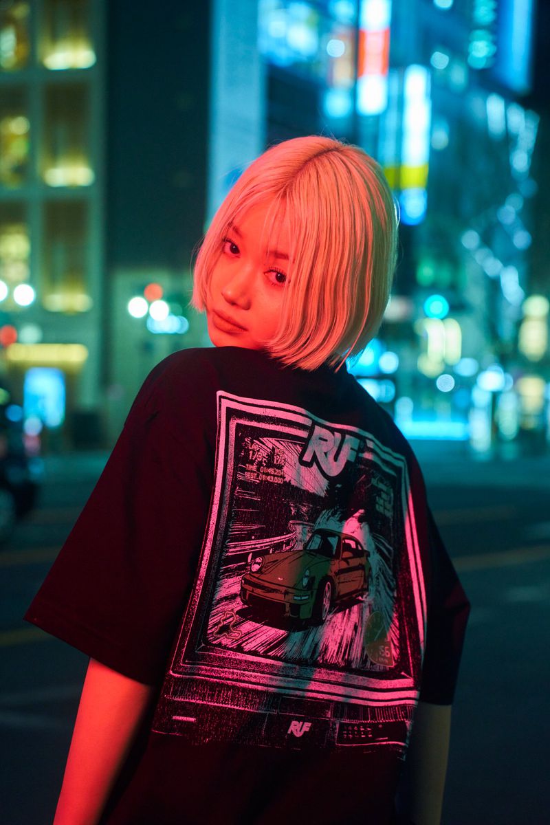 Blonde-haired woman in a black graphic tee with a car design, standing in a neon-lit city street at night, looking back at the viewer.