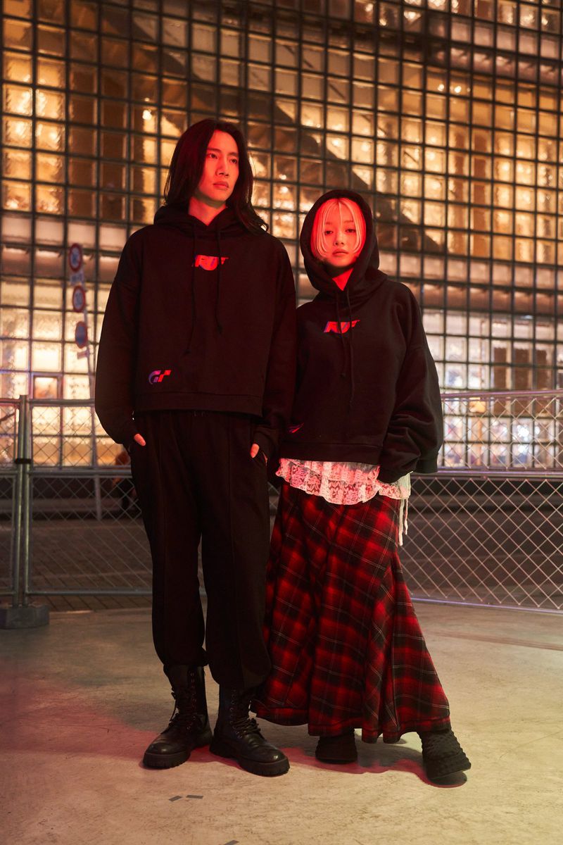 Two models wearing black hoodies and dark clothing standing outdoors at night in front of an illuminated building facade.