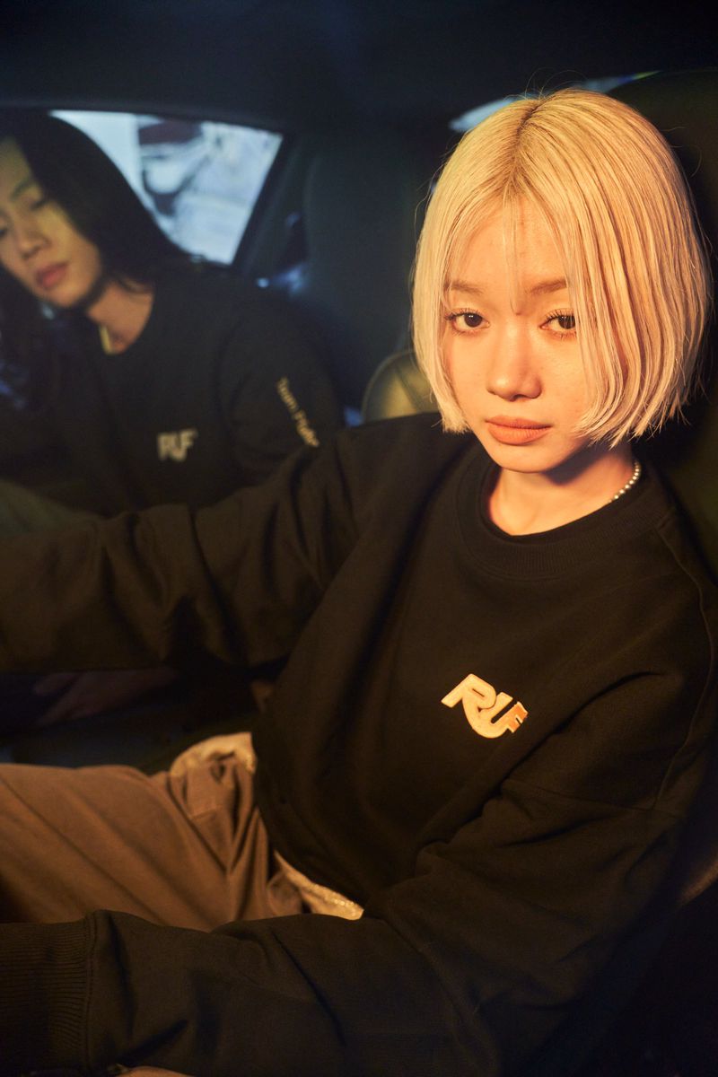 Two people sitting inside a car at night, featuring a blonde woman in a black sweatshirt in the foreground.