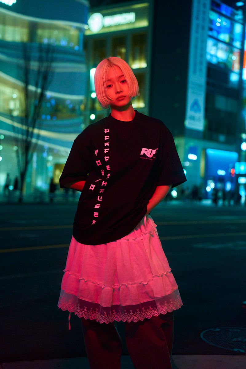 Blonde woman wearing a black graphic t-shirt and white skirt standing on a city street at night under neon lighting.
