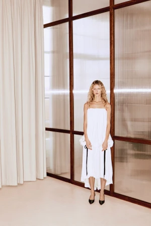 Model in white dress standing before a dark-framed glass wall and cream curtains
