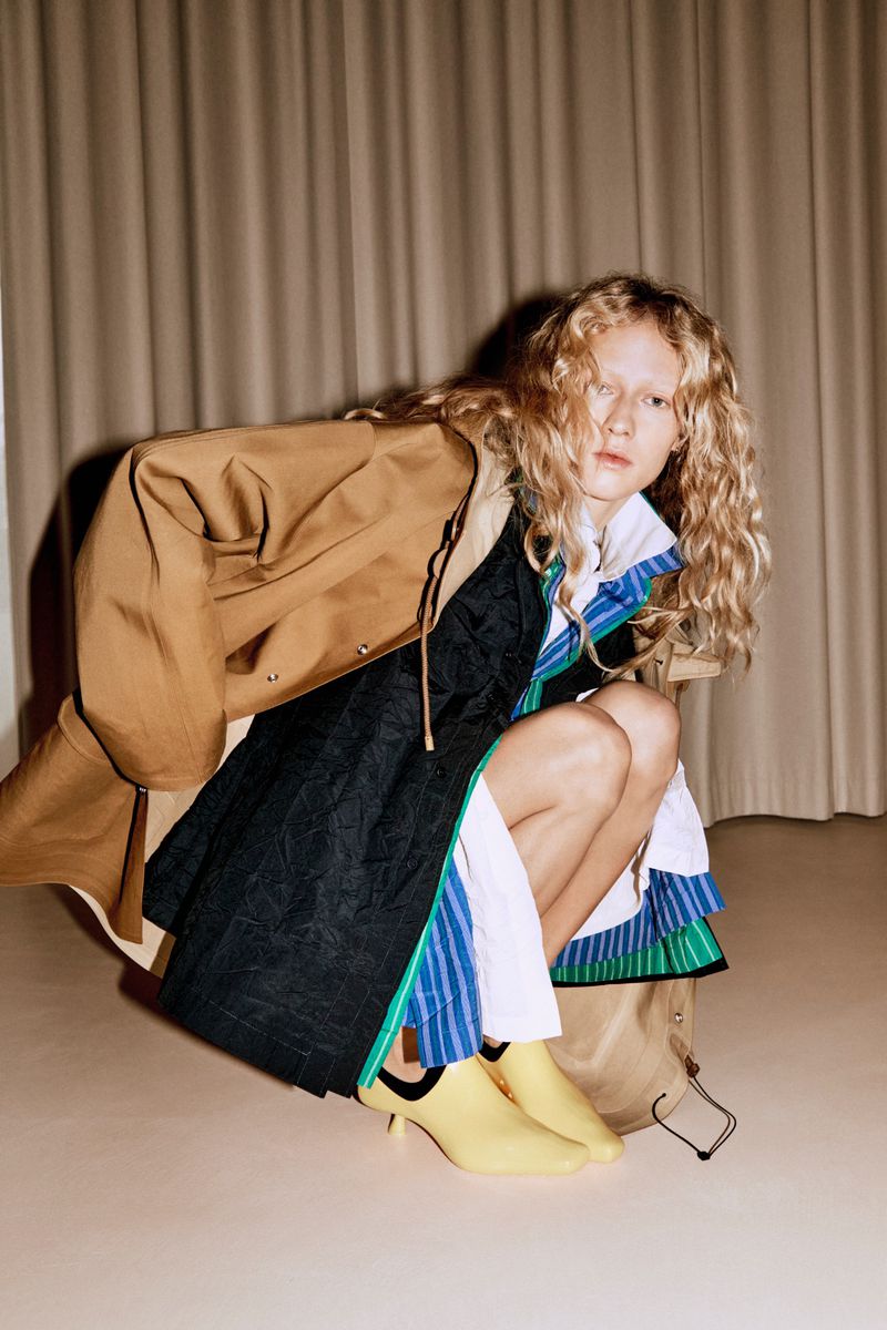 Model with long curly blonde hair wearing a camel coat and layered dress crouching against draped curtains