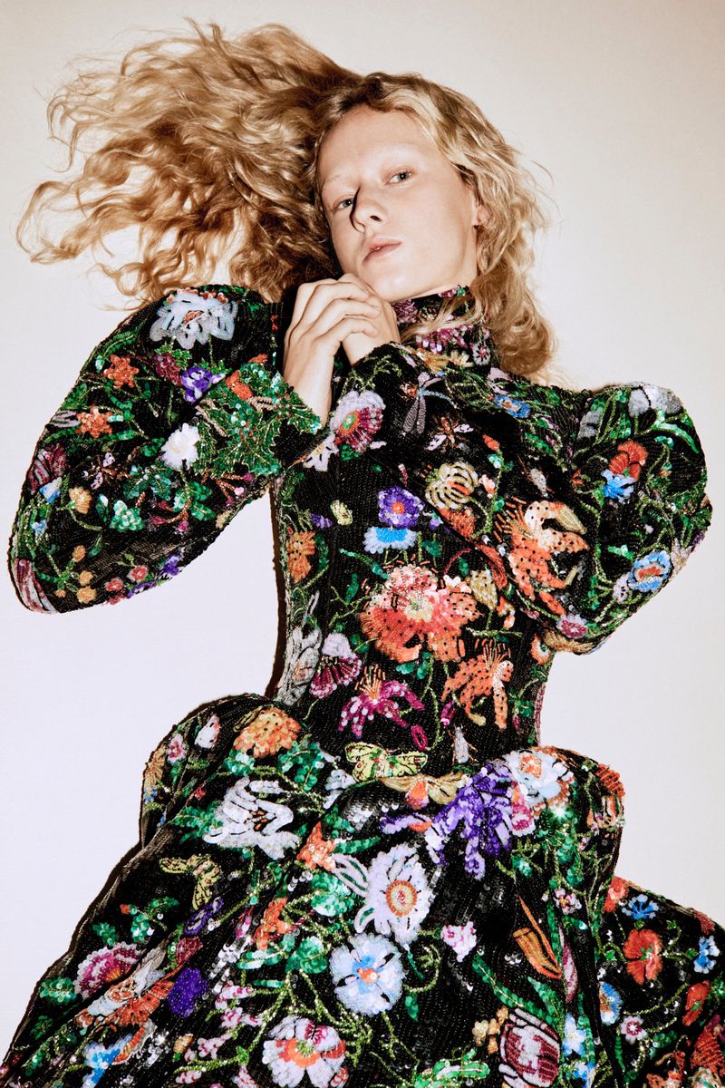Model with long blonde curly hair wearing a black dress embroidered with colorful floral patterns against a plain background