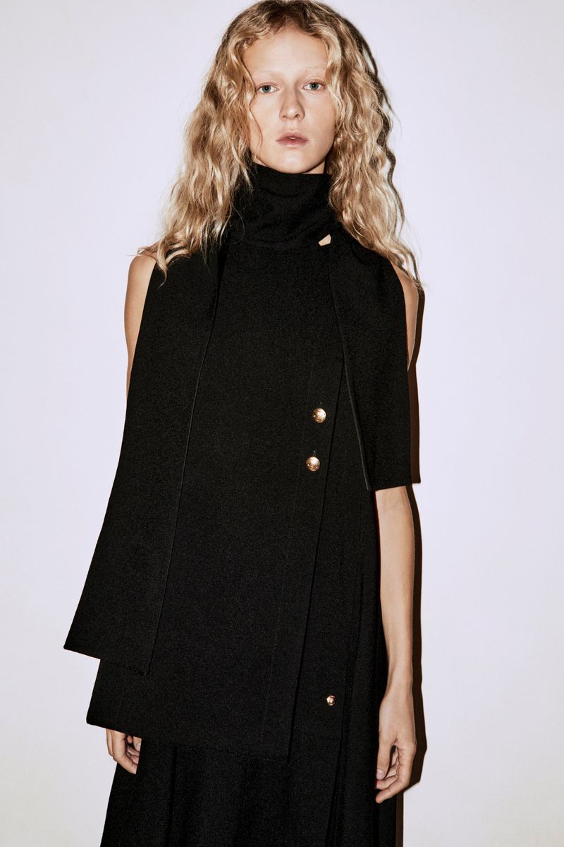 Model with long blonde curly hair wearing a black sleeveless dress and turtleneck against a plain white studio background.