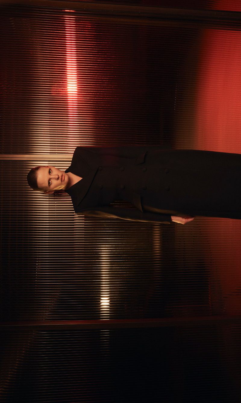 Model Lara Stone in a black coat against a textured ribbed glass background with red lighting