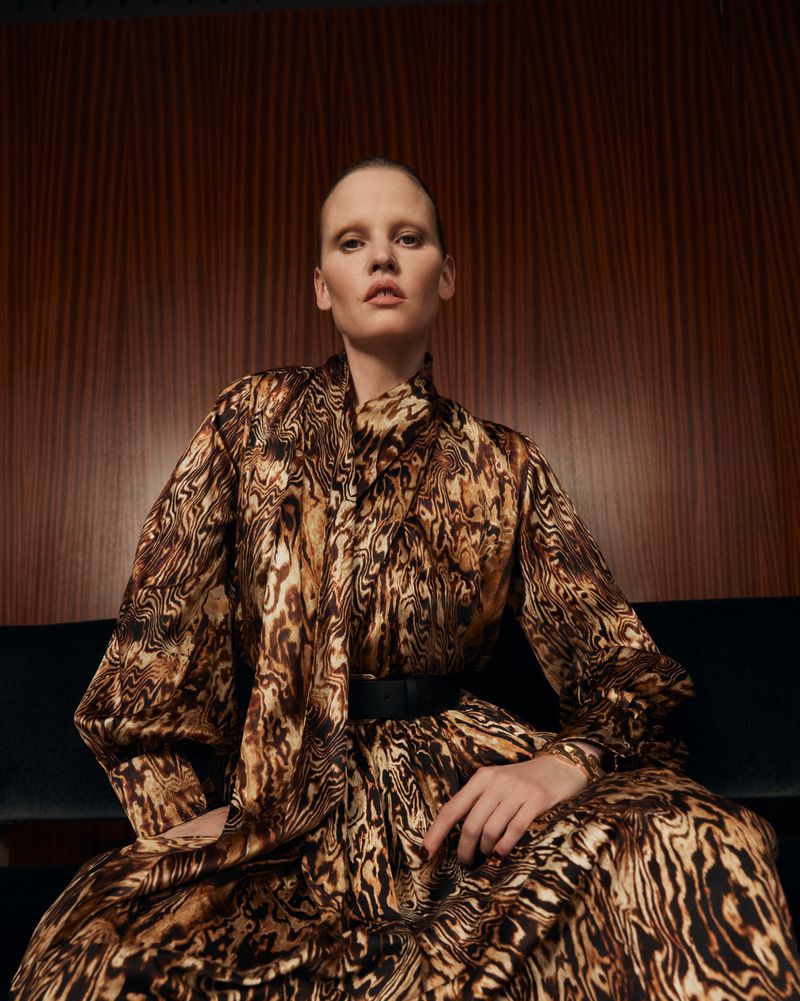 Fashion model in an animal print dress with a black belt poses against a dark wood wall, looking directly at the camera.
