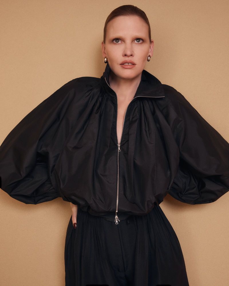 Model Lara Stone wearing a black zip-up jacket against a neutral tan studio backdrop for the Adam Lippes Spring 2026 Campaign.