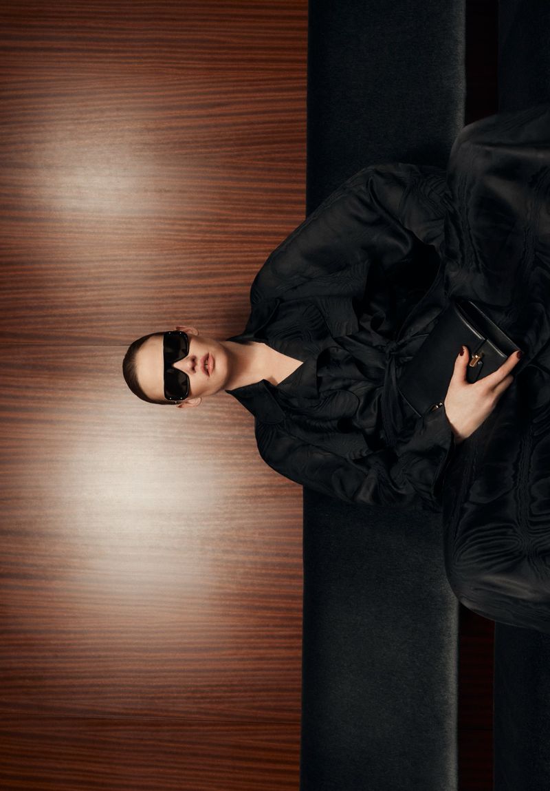 Model Lara Stone wearing a black outfit and sunglasses holding a black handbag against a wood panel wall