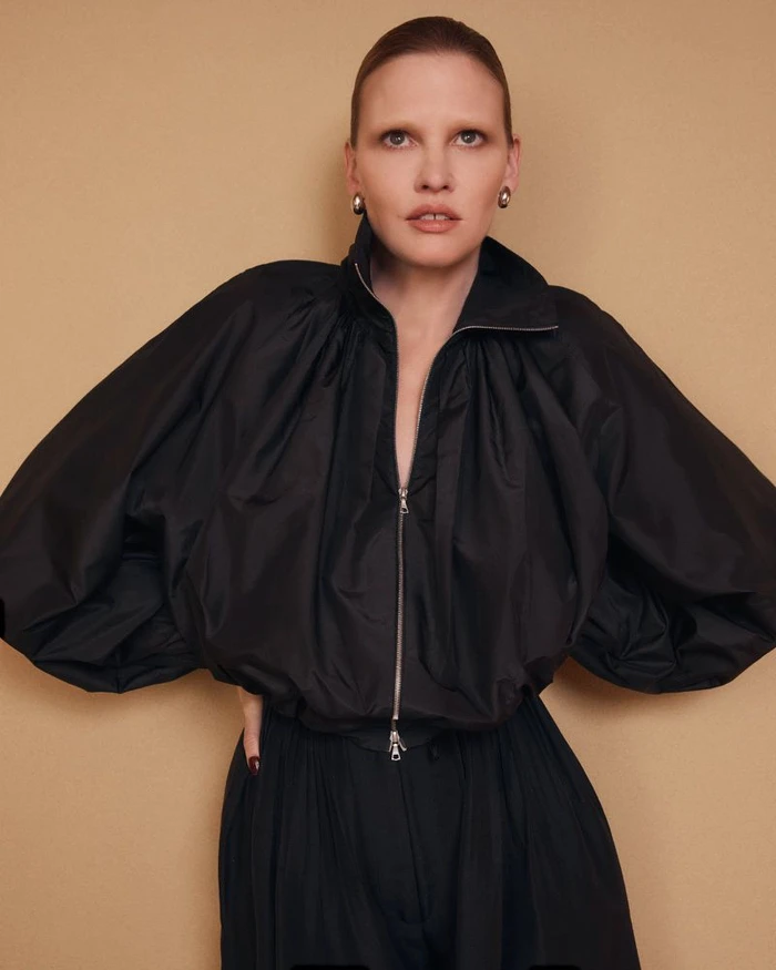 Model Lara Stone wearing a black zip-up jacket against a neutral tan studio backdrop for the Adam Lippes Spring 2026 Campaign.