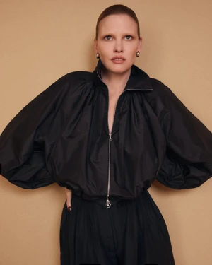 Model Lara Stone wearing a black zip-up jacket against a neutral tan studio backdrop for the Adam Lippes Spring 2026 Campaign.