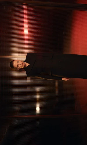 Model Lara Stone in a black coat against a textured ribbed glass background with red lighting