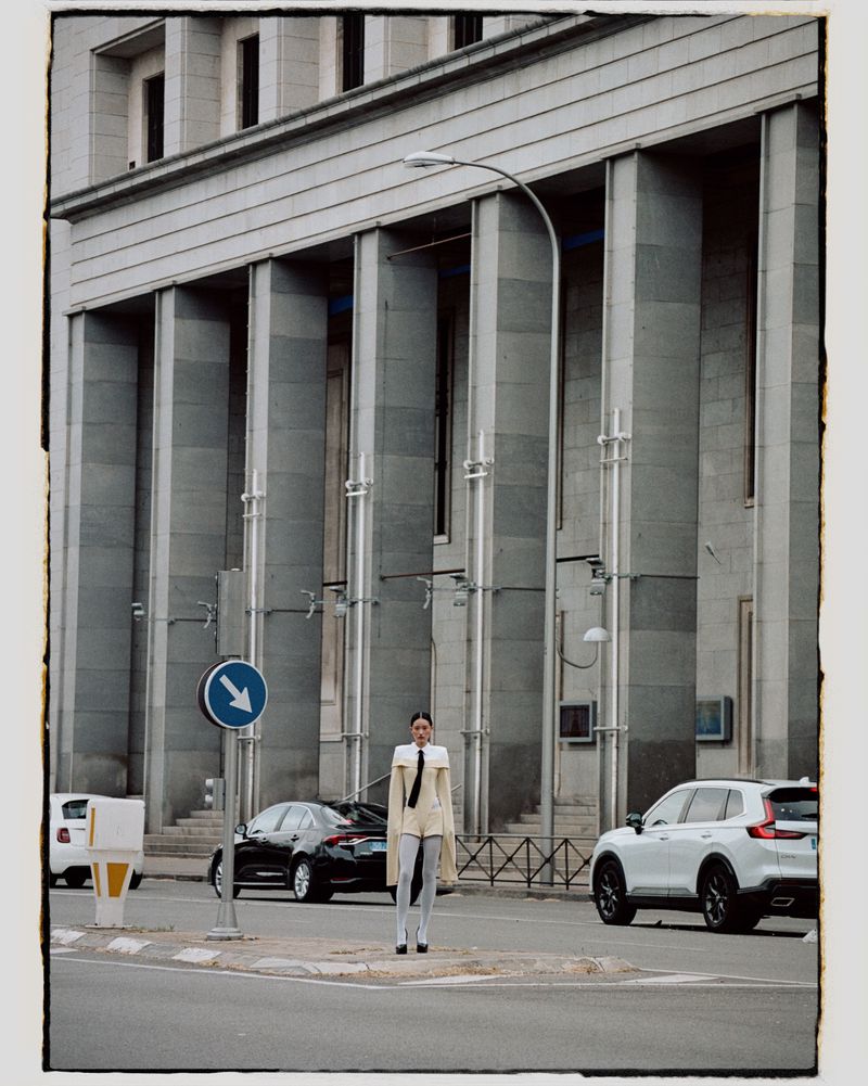 Woman in unique outfit standing on city sidewalk near intersection with cars, blue arrow sign, and grand building in the background.