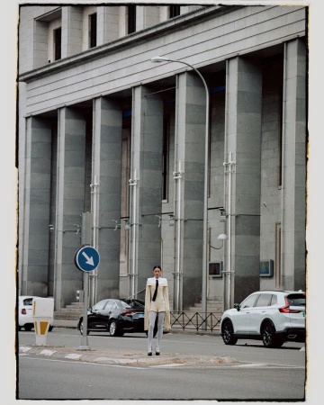Woman in unique outfit standing on city sidewalk near intersection with cars, blue arrow sign, and grand building in the background.