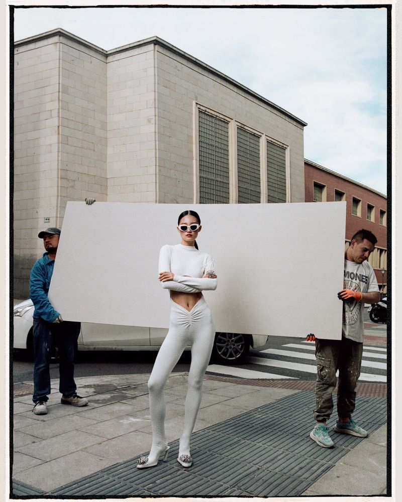 Fashion shoot featuring a slender model in an all-white outfit, posing on a city street with men holding a white backdrop.