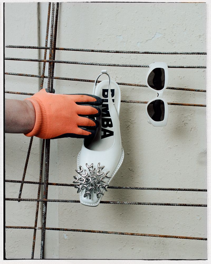 Still life of a white, embellished Bimba shoe held by a gloved hand next to sunglasses on a metal rack against a concrete wall.