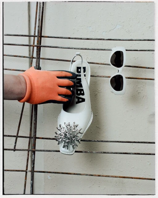 Still life of a white, embellished Bimba shoe held by a gloved hand next to sunglasses on a metal rack against a concrete wall.