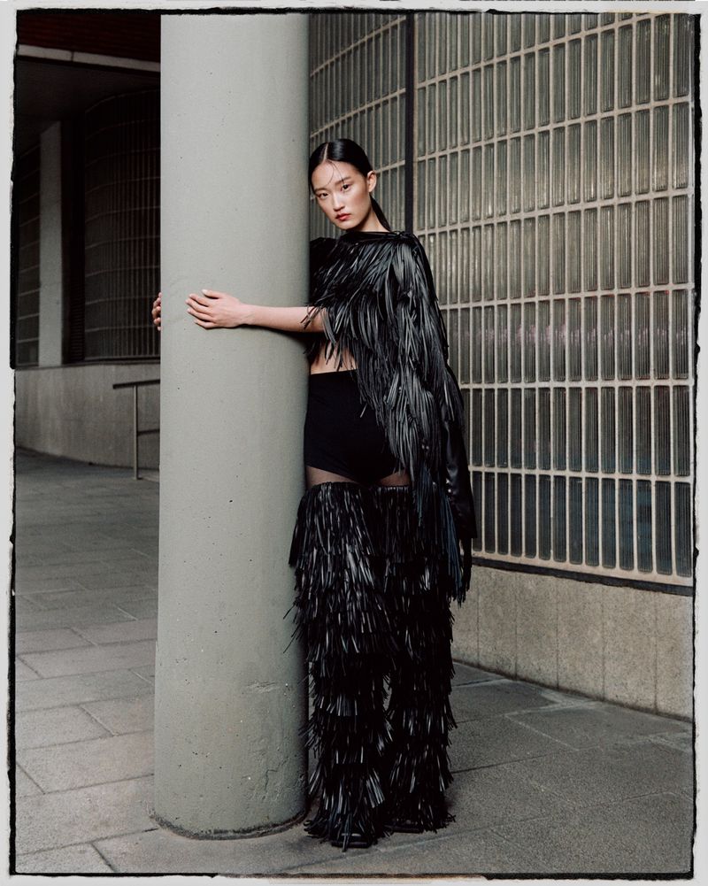An Asian woman in black fringed couture poses against a concrete pillar, wearing a black crop top and skirt with architectural background.