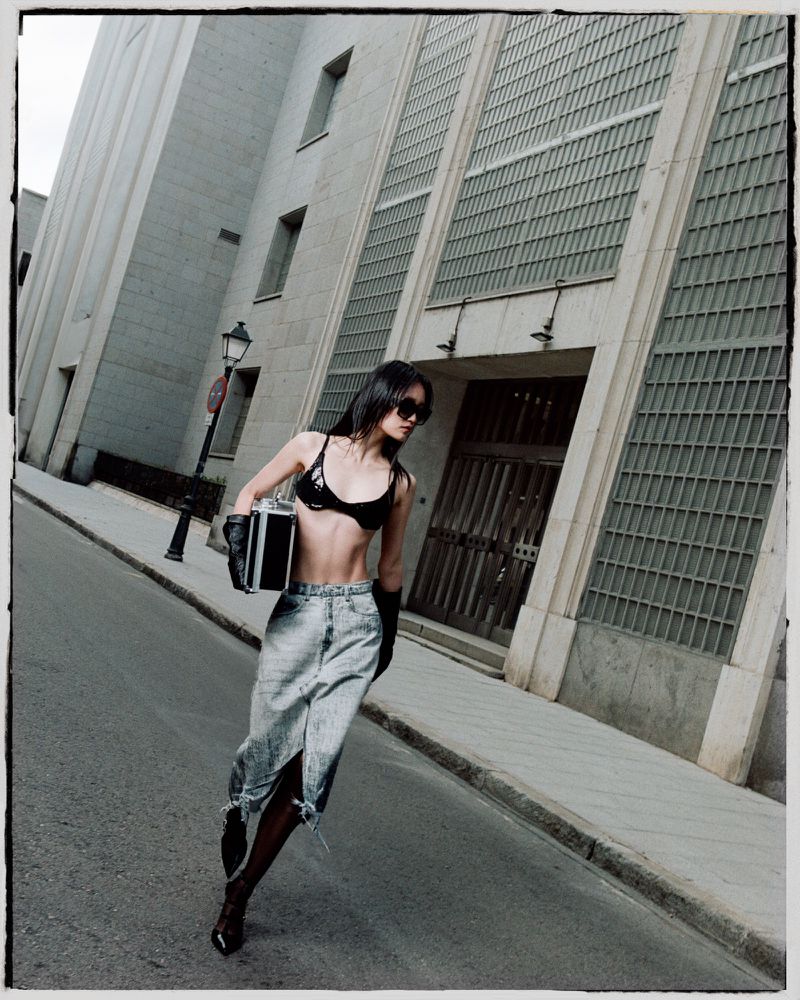 An adult woman in a black bikini top and light-wash jeans walks on a city street with a briefcase, next to a large modern building.